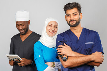 Fototapeta premium Portrait of positive healthcare workers standing in the hallway and looking at the camera. Group of medics smiling over gray background. muslim doctor woman on background of african and indian doctor