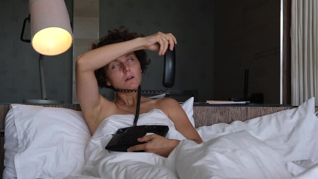 Quarantine in a hotel room during covid-19 pandemic outbreak, travel restrictions. Exhausted tired woman playing and choking herself with wire of dial phone, laying in the white hotel bed