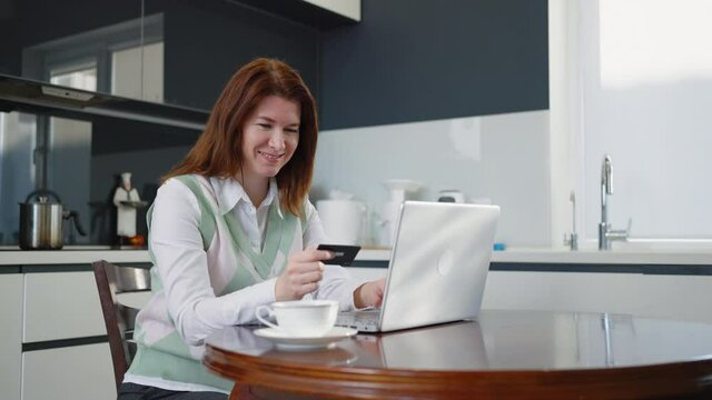Smiling woman holding credit card and entering numbers on laptop, making online order while sitting in kitchen, making yes gesture and thumb up. Concept of online shopping