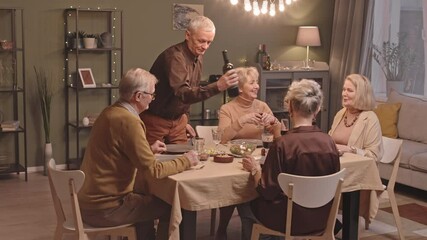 Five cheerful Caucasian seniors having dinner party together at cozy living room with lights. Man pouring red wine in glasses and making toast - Powered by Adobe
