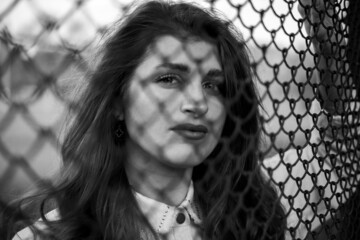 the girl looks at the camera through the net. look through the fence. model on the other side of the fence. black and white photography. out of focus people.