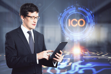 Attractive young european businessman using tablet with glowing ICO hologram on blurry office interior background. Initial coin offerng and cryptocurrency concept. Double exposure.