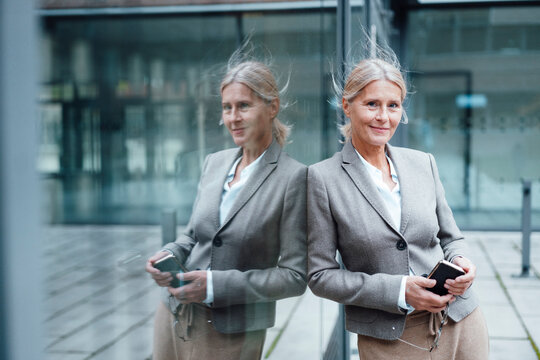 Smiling businesswoman with mobile phone leaning on glass wall