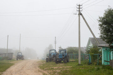 Fototapeta na wymiar Misty morning in the countryside. View of the village street. Old agricultural tractors on the side of a dirt road. Lonely man in the fog. Foggy rural landscape. Everyday village life. Russia.