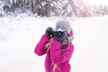 Backlit portrait of girl in warm clothes taking photo in winter forest. Child with camera for walk in winter. Day of photographer. Hobby, photography school. Lens looking at camera.