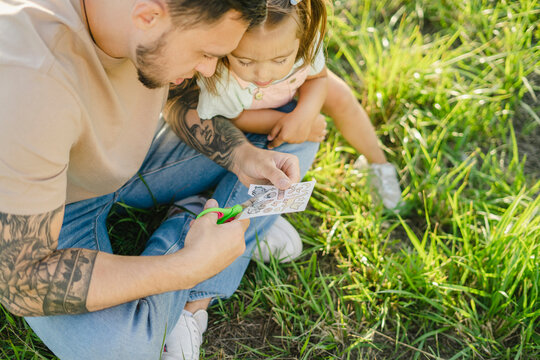 Curious Girl Sitting By Father Cutting Transferable Tattoos On Grass