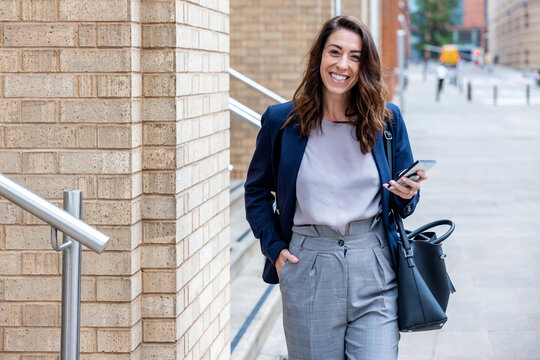 Smiling businesswoman with hand in pocket holding mobile phone walking on footpath