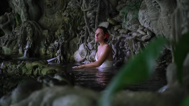 Beautiful woman with red flower in hair meditating and bathing in Bali water pond with Indonesian sculptures in tropics.