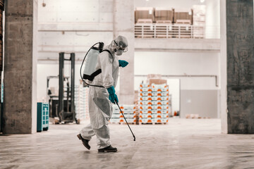 The serious situation with the coronavirus. Suppression of COVID19 and cleaning place in factory warehouse by a person in a protective white suit. Cleaning sprayer, stay protective, corona situation