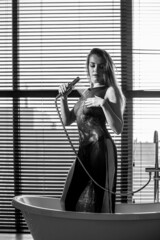 A young, slender girl in the bathroom stands naked against the background of a window with blinds.