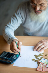 Old bearded senior man with calculator and bills counting euro money and writing notes on white sheet of paper.