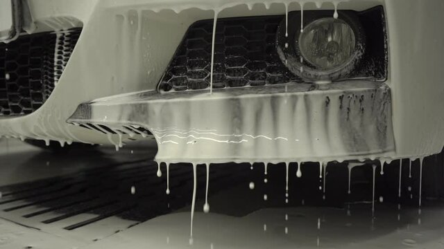 Car washing process of luxury car full covered with white foam and splashes from water gun. Spreading of foam on shiny car exterior slow motion.