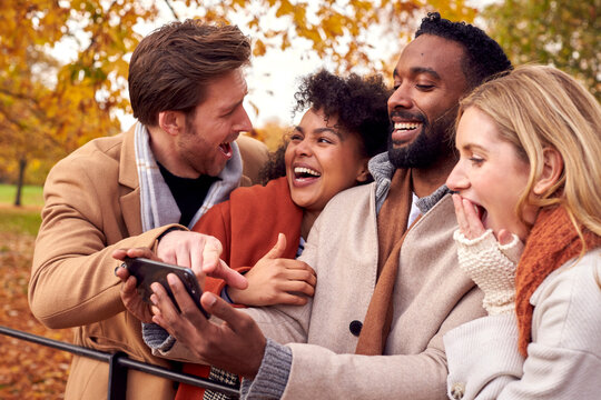 Group Of Friends Outdoors Wearing Coats And Scarves Looking At Photos On Phone In Autumn Park