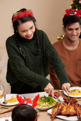 Positive young woman cutting fried turkey fro guests at Christmas dinner