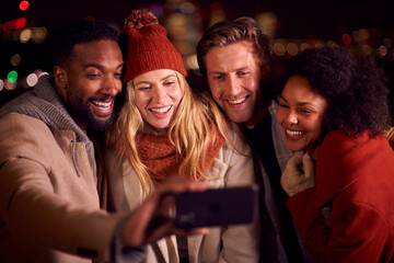Group Of Friends Outdoors Wearing Coats And Scarves Posing For Selfie On Phone Against City Lights