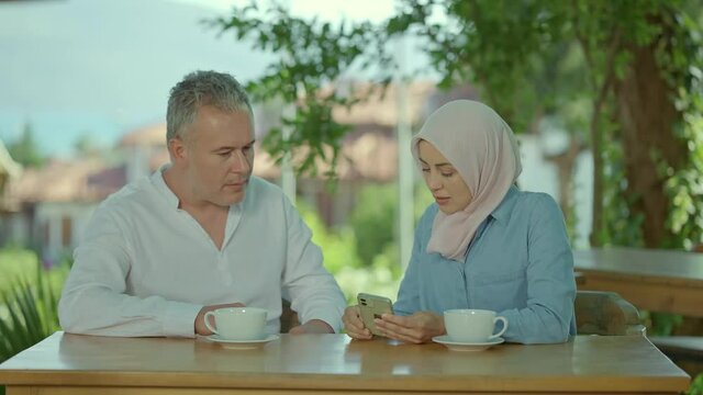 The woman gets angry by showing her husband the message or photo she sees on her smartphone.Problem in marriage, infidelity concept.Young woman in hijab and her husband drinking tea or coffee in cafe.