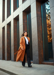 Asian business woman in long beige coat and hat walks down modern street on background of building in urban style