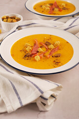 Pumpkin cream soup with crackers and jerky meat chips in a white plate on a light marble background with textiles, top view. Vertical orientation, close-up, no people