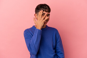 Young mixed race man isolated on pink background blink at the camera through fingers, embarrassed covering face.