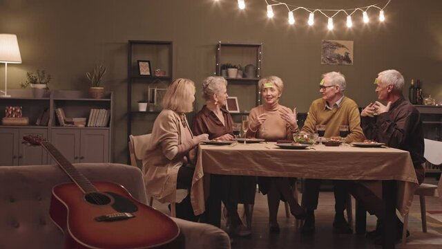 Slowmo shot of five joyful senior friends playing guessing game and toasting glasses sitting at festive dinner table in cozy living room decorated with lights