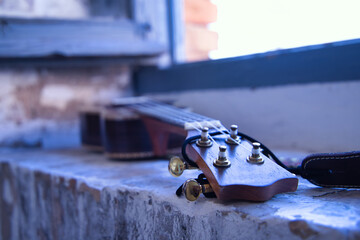 Detail of the pegbox, fret and strings of a brown ukulele with strap leaning against a brick window...