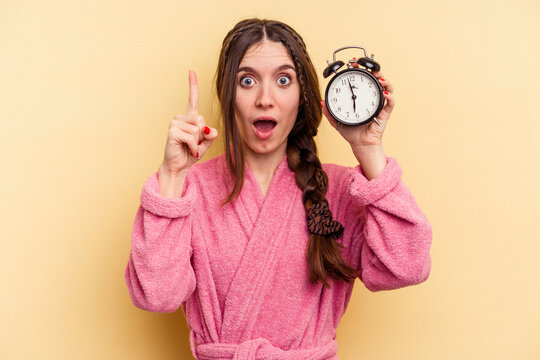 Young caucasian woman wearing a bathrobe holding a alarm clock isolated on yellow background having an idea, inspiration concept.