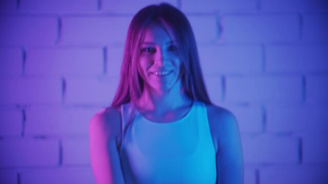 Smiling young woman exercising in neon lighting - using small dumbbells and looks in the camera