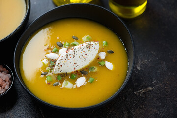 Pumpkin soup topped with soft cheese, scallions and pumpkin seeds in a black bowl, middle close-up, studio shot
