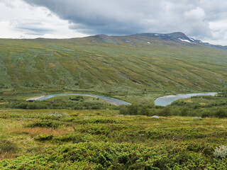 Obraz premium Valley with winding Laddejahka river, mountain hut STF Laddejahka fjallstuga cabin, birch tree forest and green mountains. Lapland landscape Sweden at Padjelantaleden hiking trail. Summer moody sky
