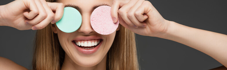smiling woman obscuring eyes with cosmetic sponges isolated on grey, banner