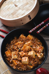 Black bowl with chinese traditional mapo tofu, vertical shot with a bamboo steamer in the background