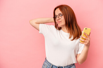Young caucasian woman holding a mobile phone isolated on pink background touching back of head, thinking and making a choice.