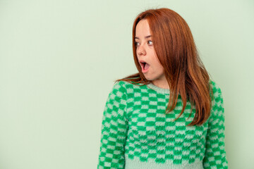 Young caucasian woman isolated on green background being shocked because of something she has seen.