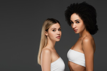 young multiethnic women in white tops and natural makeup looking at camera isolated on grey