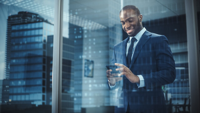 Portrait of Successful Black Businessman Wearing Suit Standing, Using Smartphone Looking out of the Window. Successful African CEO Planning e-Commerce Investment Strategy. From Outside Shot