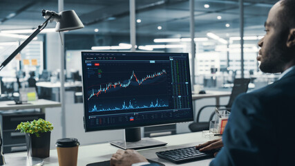 Successful Black Businessman Sitting at Desk Working on Desktop Computer in Big City Office. Hard Working Top Manager Doing Research for e-Commerce Project with Graps, Charts and Tables. Over Shoulder