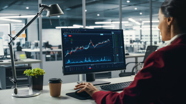 Successful Business Woman Sitting at Desk Working on Desktop Computer in City Office. Hard Working Top Manager Doing Research for e-Commerce Project with Graps, Charts and Tables. Over Shoulder
