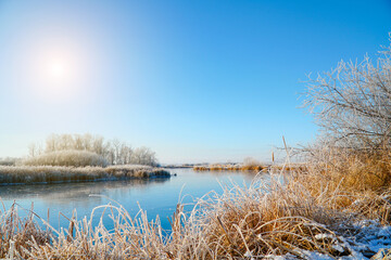 The sun is over the ice of the winter small river. Bushes, reeds and trees covered with frost. Fabulous Winter landscape
