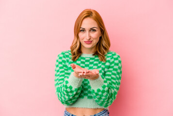 Young caucasian woman isolated on pink background holding something with palms, offering to camera.