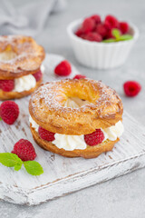 Choux pastries. Choux rings with cream of cream cheese or cottage cheese and fresh raspberries, dusted with powdered sugar on a wooden board on a gray concrete background.