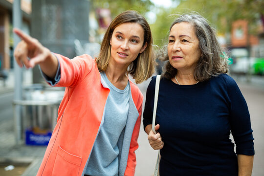 Friendly girl helping elderly woman to find necessary address in city, pointing direction with hand.
