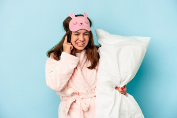 Little caucasian girl going to sleep isolated on blue background covering ears with hands.