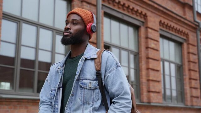 Listening popular music at walking. Black guy student with wireless earphones puts phone into pocket and presses key on pad track by street building spbas
