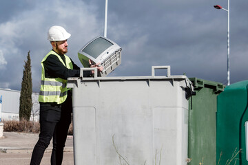 Trabajador del centro de reciclaje arrojando un viejo televisor de tubo en un contenedor de basura gris. Hombre que lleva un chaleco reflectante y casco protector. Reciclaje de aparatos electrónicos