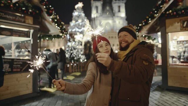 Lovers travelling to Europe at the height of Christmas holidays. Sweethearts kissing in the centre of old city. Romantic couple in love hugging and burning the sparklers at festive street fair.
