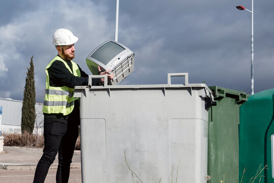 Trabajador del centro de reciclaje arrojando un viejo televisor de tubo en un contenedor de basura gris. Hombre que lleva un chaleco reflectante y casco protector. Reciclaje de aparatos electr&oacute;nicos