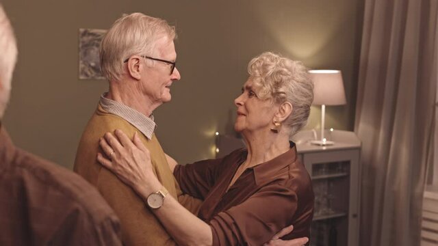 Medium slowmo shot of happy Caucasian senior couple slow dancing in cozy living room during home party with friends