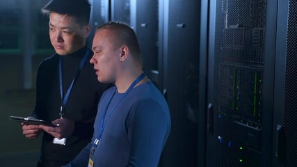 Two male engineers talking and checking equipment while standing in data center spbas. Close-up view of young men examine hardware racks and talk, use tablet in hands and stand in dark database