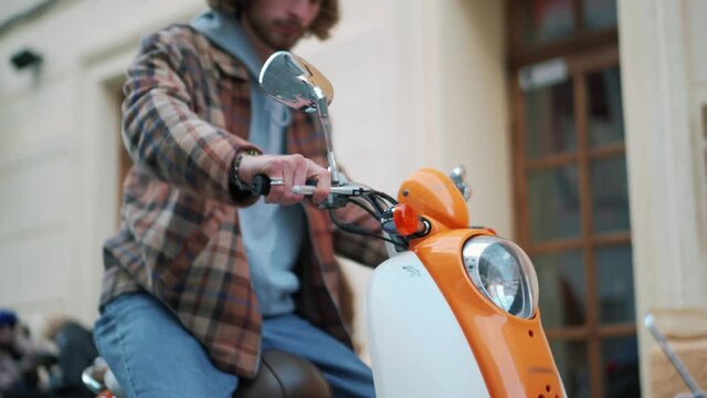 Concentrated curly-haired man gets on a moped and leaves on the street