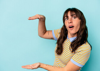 Young Argentinian woman isolated on blue background shocked and amazed holding a copy space between hands.